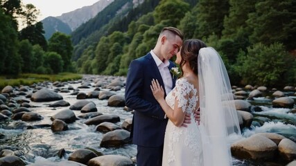 Romantic matrimonial partners sharing intimate gesture along rocky waterway edge in scenic alpine setting