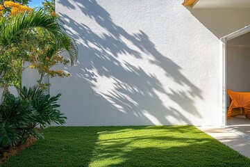 Sunny outdoor patio with lush green grass and tropical plants casting shadows on a white wall beside an open door with orange woven chairs inside