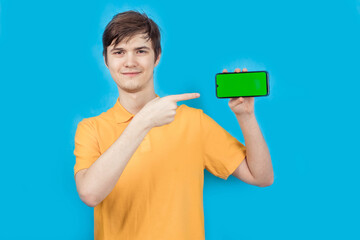 Young man, on a blue background, holding a smartphone with a green screen in his hands, with his...