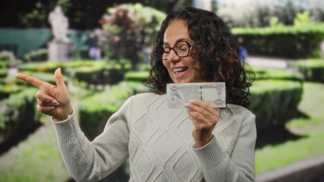 Middle-aged woman in a park pointing with excitement while holding a uae dirham banknote.