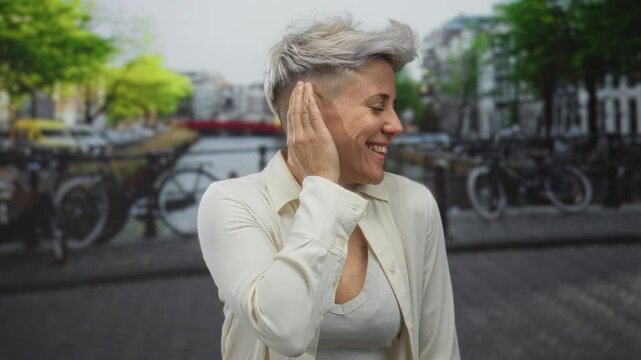 Woman with short hair touches ear by canal street wearing white shirt and tank top in city; serenity introspection insight.
