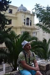 Vertical street city photo. Mature fat white man with stubble, floral print beach shirt, hat, blue shorts, sits on remains of stone old fortress wall and looks at side. Summer vacation, rest, trip