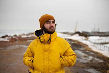 Horizontal travel photo. Male portrait. Mature white man with beard, yellow winter jacket with hood, brown hat stands on snowy sea coast. Ocean beach. Cold winter weather. Tourism, adventure, trip