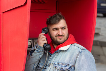 Horizontal street city photo. Mature fat white man in blue denim jeans jacket, red hoodie talks from stationary payphone in retro vintage telephone booth. Concept of public phone, call, trip, travel