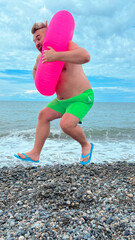 Mobile vertical photo. Mature fat white man with big belly, bright green shorts, pink magenta swim ring and blue flip-flops jumps on sea pebble coast. Concept of summer trip, rest, calm, travel
