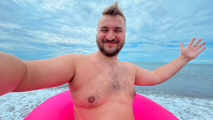 Horizontal mobile selfie travel photo. Mature tanned fat white man with beard, big belly, pink swim ring on pebble sea beach looks and smiles at camera. Sun day. Blue sky. Summer vacation, trip, walk