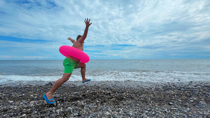 Mobile wide angle travel photo. Back view. Mature fat white man with big belly, bright green shorts, pink swim ring, flip-flops runs and jumps to sea on pebble beach. Blue sky. Summer trip, rest, calm