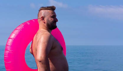 Horizontal travel photo. Mature fat white naked man with beard, big belly, magenta inflatable swim ring on sea shore, coast. Blue sky. Sun day. Concept of tourism, summer rest, trip, relax. Copy space