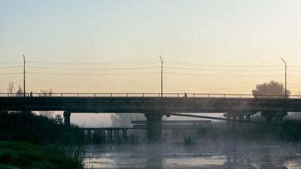 Misty bridge at sunrise with cyclists over calm river