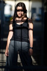 Teenager posing on urban street. Stylish teen girl with confident look. Young female model. Hipster girl standing in shadows. Gen Z fashion portrait outdoors. Trendy teenager with serious expression.