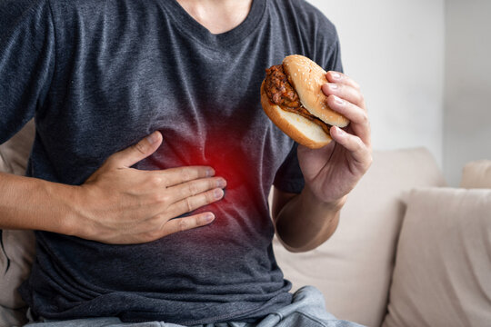 Asian Man Suffering from Stomach Pain and Discomfort After Eating Junk Food, Holding a Burger in Hand Experiencing Heartburn and GERD Symptoms