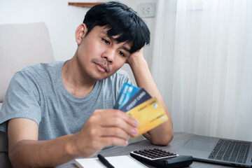 Stressed Asian Man Sitting at Home Holding a Credit Card, Struggling with Debt and Financial Issues...