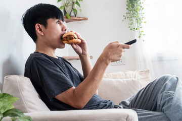 Asian Man Having a Lazy Day on the Sofa, Eating Fast Food burger While Watching TV, Avoiding...