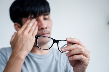 Tired Asian Man Experiencing Eye Pain and Blurry Vision, Holding His Eyeglasses in Hand While...