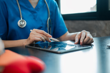 Close-up of a healthcare worker in blue scrubs using a tablet with a stylus in a medical office.