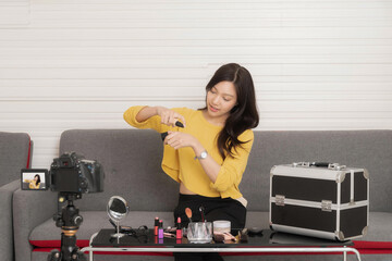 Cheerful Asian woman sits on a sofa with makeup products on a table, gesturing while recording a beauty vlog with a camera on a tripod.