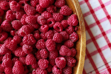 A wooden bowl with ripe raspberries lies on a rustic tablecloth.