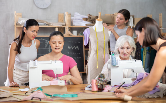 Friendly diverse group of female fashion designer students laughing and engaging while sewing on machines, handling fabric, and fitting dress on mannequin together in class - Powered by Adobe