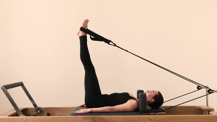 Slim brunette woman in black tight sportswear working out on Pilates reformer machine. Stretching and toning muscles during Pilates session in bright fitness studio. - Powered by Adobe