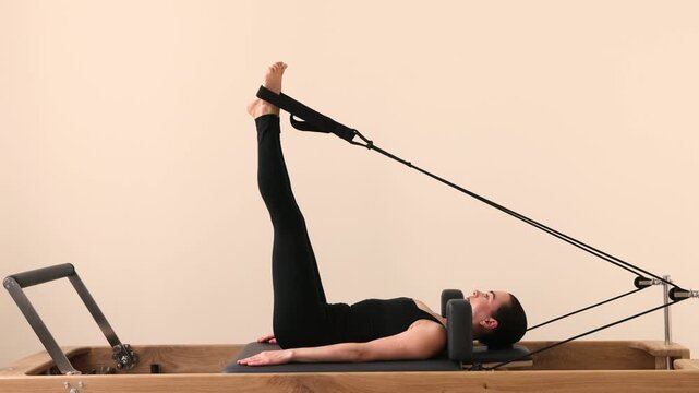 Slim brunette woman in black tight sportswear working out on Pilates reformer machine. Stretching and toning muscles during Pilates session in bright fitness studio.
