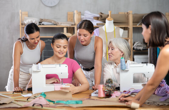 Group of interested women of different ages gathering around sewing machine in training workshop while female seamstress explaining stitching techniques and sharing professional tips..