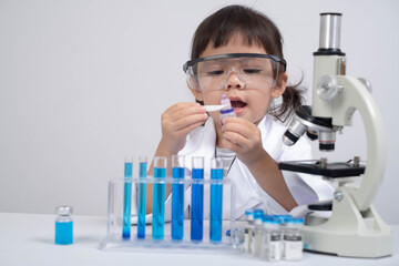 A young child in a lab coat examines a test tube while using a microscope, showcasing curiosity and...