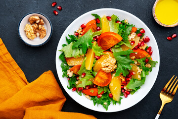 Autumn vegan persimmon salad with pomegranate seeds, walnuts, orange slices and lettuce, black background, top view