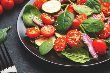 Healthy vegan salad with spinach, tomatoes, cucumber, onion, sesame and olive oil. Black table background, top view