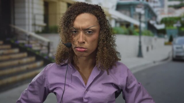 Woman grimaces with teary eyes and clenched fists on street near building wearing headset microphone for a comedic crying pose; sadness.