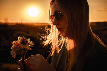 Teen girl with long blonde hair and heart-shaped sunglasses holding wildflowers in golden sunset light, enjoying a calm summer evening outdoors