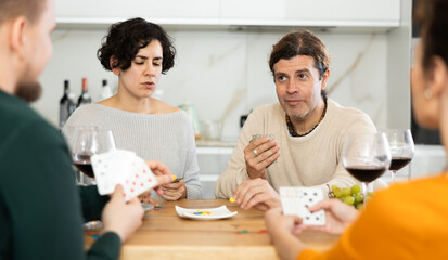Two married couples are having fun in cozy home atmosphere, playing card game poker. Player makes...
