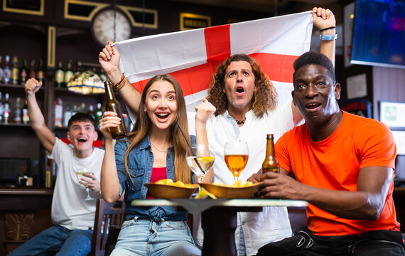 Group of cheerful football fans rooting for England team together in sports bar, waving national flag while watching match on TV