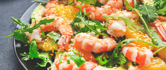 Gourmet seafood salad with shrimps, avocado, orange, arugula and sesame seeds on black background. Top view