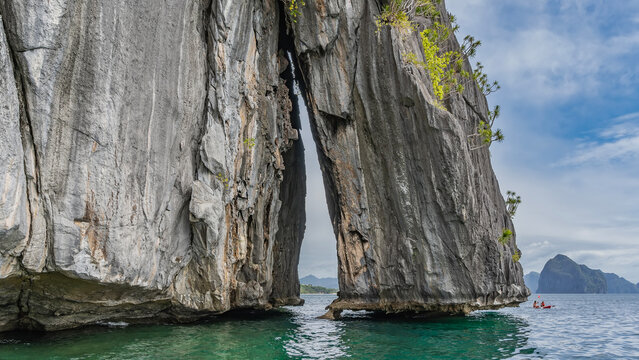 The rock rises above the ocean. There is scant green vegetation on the steep slopes. The narrow gap between the cliffs. A tourist canoe floats nearby. Mountains in the distance. Blue sky, clouds.