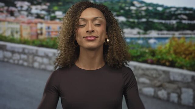 Woman flexes biceps wearing fitted top on sunlit street by stone wall; empowerment confidence pride strength.