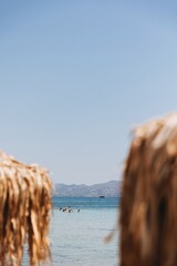 Blurry foreground of straw beach umbrellas framing a clear blue sea at Skala Beach on Agistri Island, Greece. In the background, swimmers cool off in the shimmering water on a hot summer day