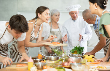 Positive interested young woman participating group culinary class, engrossed in cooking process talking friendly to other participants..
