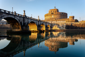 Castel Sant'Angelo and Ponte Sant'Angelo in Rome, Italy, beautifully reflected on the calm surface of the Tiber River during golden hour. This landscape photograph captures the historical architecture