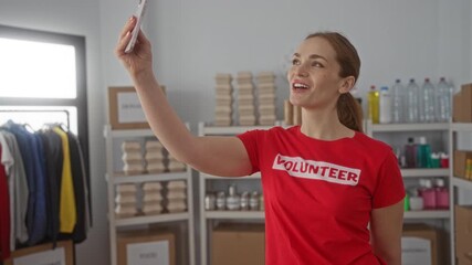 Young woman in red tshirt holds smartphone at arm’s length for a selfie in a donation center building; charity compassion.