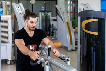 Young good looking physical trainer shows how to build upper part body by pulling up an equipment in a fitness room. A muscular sport man taking an exercise with fitness equipment to build his body.