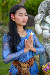 Balinese woman in blue lace kebaya kneeling in prayer beside a stone statue. Holding a frangipani flower with hands in namaste position