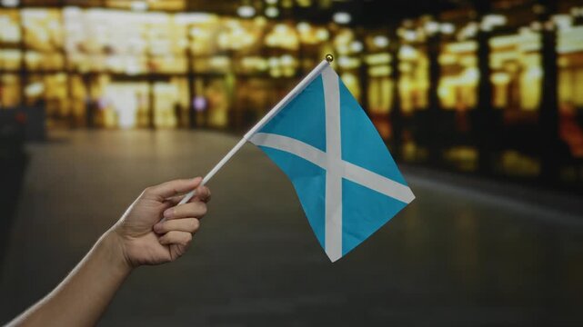 Hand holding scottish flag in city street showcasing national pride outdoors at night with blurred lights in the background.