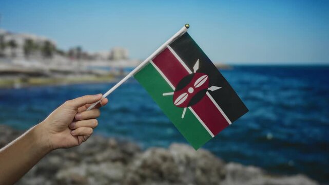Hand of caucasian man holding kenyan flag at seaside beach on sunny day.