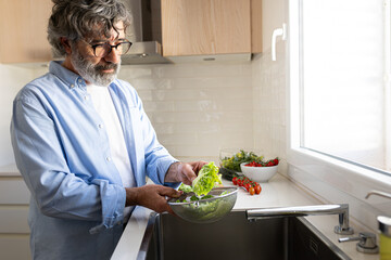 Mature man washing lettuce and vegetables in kitchen sink to prepare fresh salad for healthy lunch. Copy space.