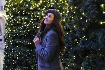 Cheerful woman in a gray coat and beret smiles while looking at glowing Christmas trees in a...