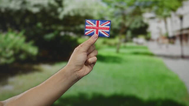 Caucasian man holding embroidered great britain flag patch outdoors in a park, showcasing patriotism amidst greenery and bright sunshine with a blurred background.