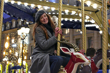 Happy woman in a gray coat and beret smiles while riding a glowing Christmas carousel at night in...