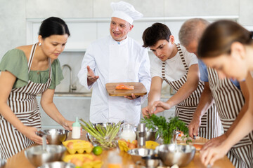 Adult male cook in uniform teaches group of different people how to cook dish