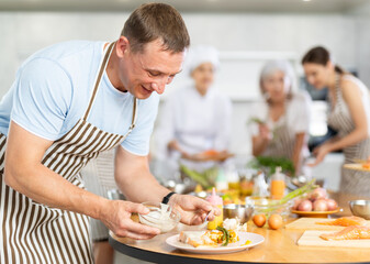 Adult man in apron pours sauce into ready dish at cooking master class