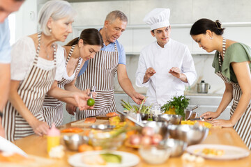 Young guy cook in uniform teaches group of different people how to cook dish